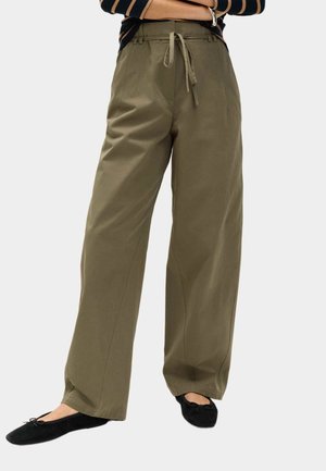 Pantaloni a gamba larga verde oliva con una texture liscia, dotati di vita con coulisse e pieghe frontali, abbinati a scarpe piatte nere.