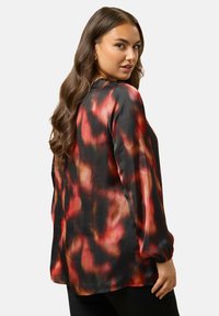 Kvinde med langt, bølget brunt hår, iført en løs bluse med lange ærmer, der har et rødt, sort og orange abstrakt mønster, mens hun ser over sin skulder.