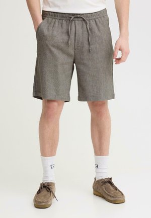 Mann trägt graue Shorts mit Kordelzug, weiße Socken und braune Wildlederschuhe, steht vor einem schlichten hellen Hintergrund.