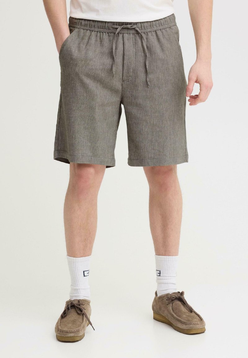Homme portant un short gris à cordon, des chaussettes blanches et des chaussures en daim marron, debout devant un fond uni clair.