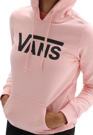 Persoon draagt een lichtroze hoodie met zwart "VANS"-logo, handen gedeeltelijk in de voorzak, en de capuchon op.