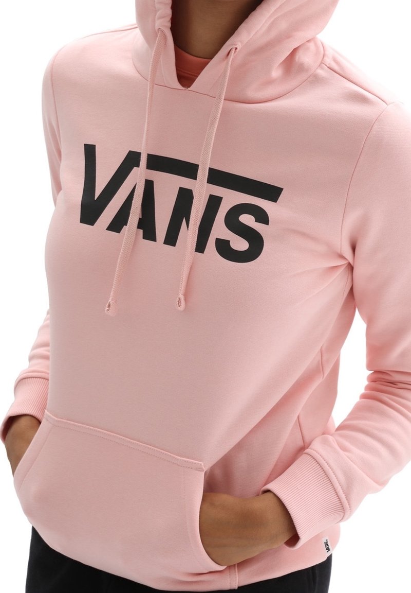 Personne portant un sweat à capuche rose clair avec un logo noir "VANS", les mains partiellement dans la poche avant et la capuche relevée.