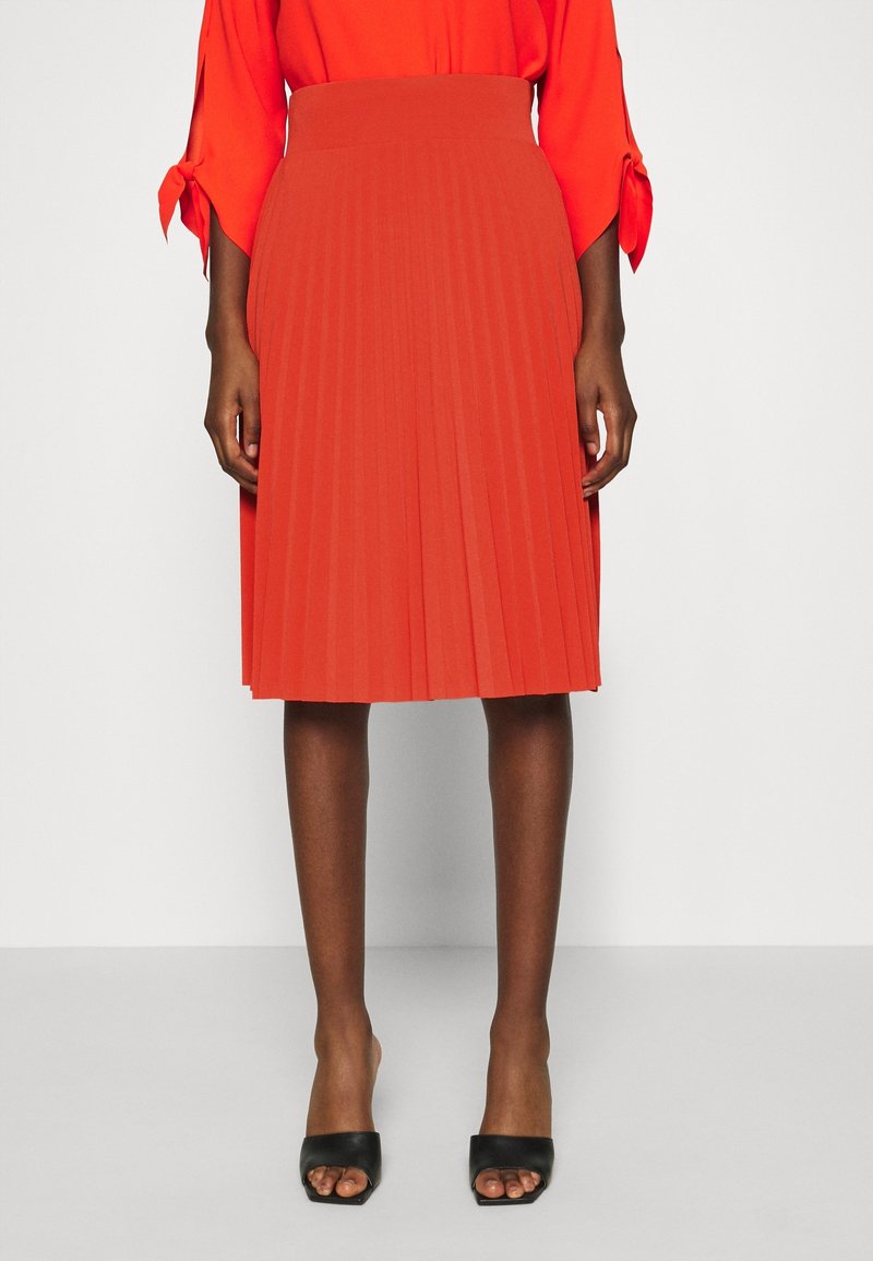 Femme portant une jupe plissée orange vif mi-longue, un haut à manches longues assorti avec des poignets noués, et des talons ouverts noirs.
