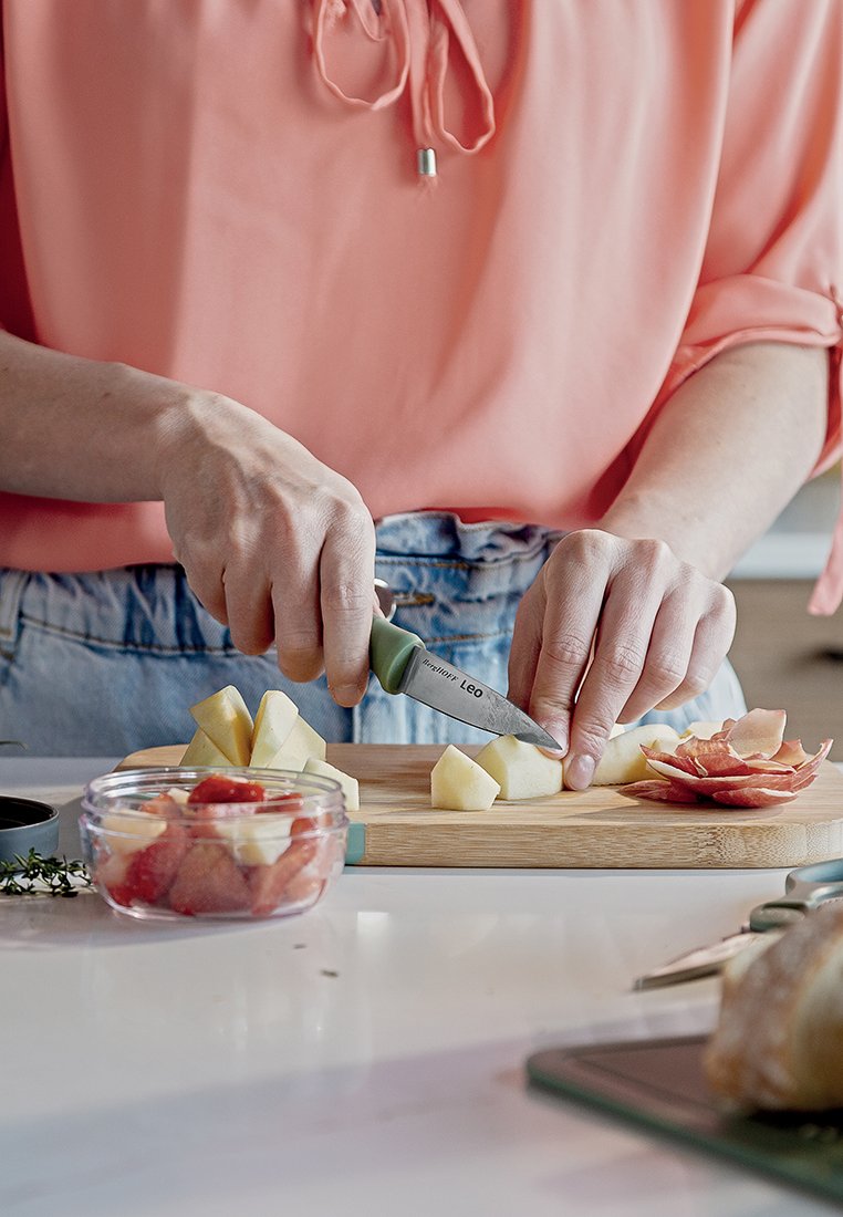 Zielono-ręcznym nożem krojone są żółte kostki jabłka na bambusowej desce do krojenia. Obok leży miska z posiekanymi truskawkami i ziołami.