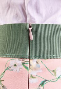 Detailní záběr na růžovou květinovou sukni se zeleným žebrovaným pasem a uzavřeným růžovým zipem uprostřed bílé látky.