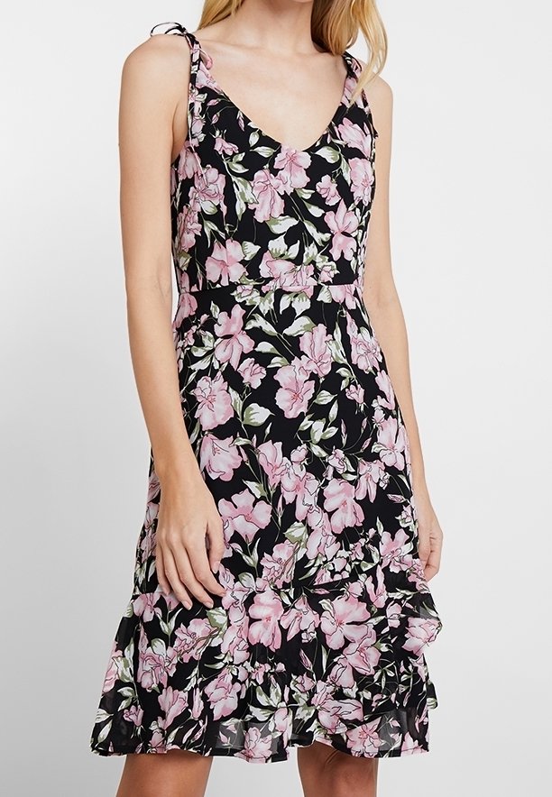 Femme portant une robe noire sans manches à motif floral rose, encolure en V, ourlet à volants et bretelles nouées, debout devant un fond uni.