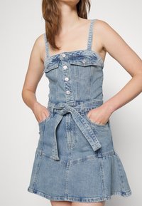 Jasnoniebieska sukienka dżinsowa z dopasowanym korpusem, kwadratowym dekoltem i detalami w postaci guzików. Posiada rozkloszowany dół oraz paskiem w talii i kieszeniami po bokach.