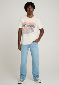 Cream-colored cotton t-shirt featuring a retro cassette design and text, paired with light blue denim jeans and navy sneakers.