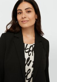 Czarny żakiet o strukturalnym kroju i klapkach, noszony na wzorzystej białej bluzce z czarnym odręcznie napisanym tekstem.