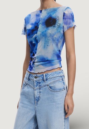 Frau trägt ein kurzärmeliges, blaues Batik-Crop-Top und hellblaue hoch taillierte Jeans aus Denim.