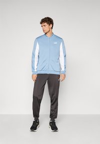Lyseblå zip-jakke med hvide detaljer, sorte træningsbukser og sorte sneakers. Materialet ser glat og sporty ud. Puma-logoet er synligt.