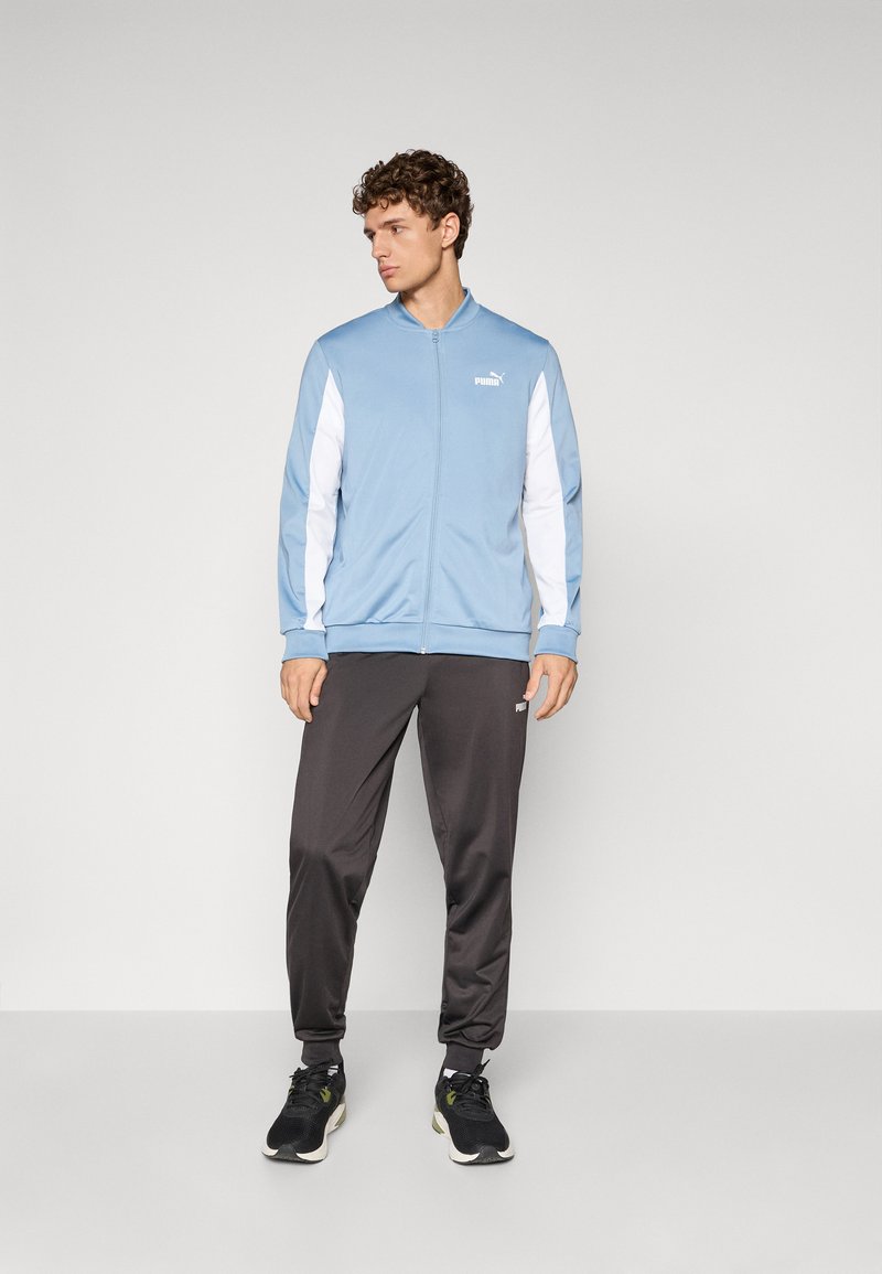 Lyseblå zip-jakke med hvide detaljer, sorte træningsbukser og sorte sneakers. Materialet ser glat og sporty ud. Puma-logoet er synligt.