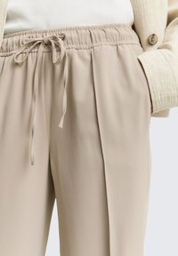 Pantalones ligeros beige con cintura elástica y detalle de cordón, que presentan una textura suave y pinzas laterales.