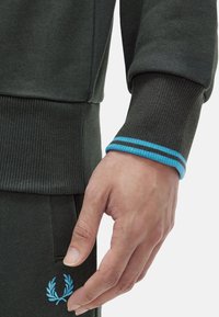 Sudadera verde oscura con textura acanalada y un puño a rayas azules, que presenta un logo azul bordado en el área del bolsillo.