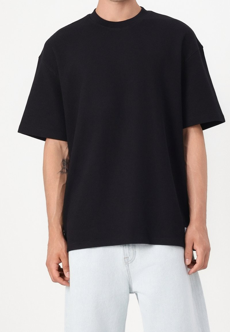 Personne portant un t-shirt noir uni oversized à manches courtes et un jean bleu clair, debout devant un fond blanc.