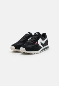 Ett par svarta och vita avslappnade sneakers med svarta snören, vit sula och en röd etikett på sidan, uppställda mot en vit bakgrund.