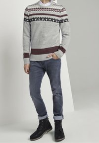 Homme portant un pull gris à motifs, un jean bleu retroussé et des bottes noires à lacets, debout avec une main dans la poche devant un fond uni.
