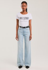 Žena oblečená v bílém tričku s nápisem "BIG STAR", zastrčeném do světle modrých širokých džín s černým opaskem a černými špičatými botami, stojící před jednobarevným pozadím.