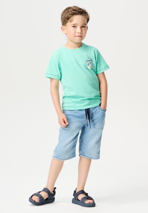 Mintgrünes Baumwoll-T-Shirt mit blauem Emblem, kombiniert mit hellblauen Denim-Shorts mit Kordelzug. Schwarze Sandalen vervollständigen das Outfit.