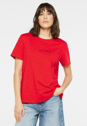 Woman with shoulder-length brown hair wearing a red T-shirt with "SENSES" text and light blue jeans, posing with one hand in pocket.