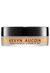 Baume de fondation dans un pot transparent avec un couvercle noir. Le produit de couleur crème est visible à l'intérieur, mettant en valeur sa texture lisse. Nom de la marque affiché.