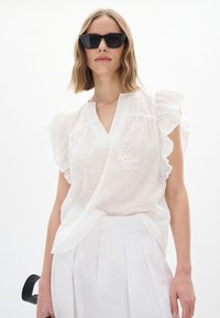 Vit blus i broderie anglaise med volangärmar, V-ringning och lätt, strukturerad tyg. I kombination med högtalande vita byxor.