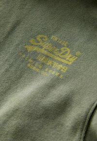 Tessuto verde oliva con una morbida texture, caratterizzato da un logo giallo "SuperDry" e ulteriore testo in stile vintage.