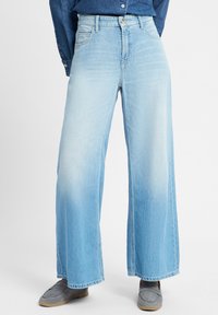 Hellblaue weit geschnittene Jeans aus Denim. Hat eine hohe Taille, einen verwaschenen Effekt und rohen Säumen. Kombiniert mit grauen Slippern.