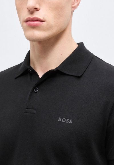 Gros plan sur une personne portant un polo noir avec un col et deux boutons, arborant le mot "BOSS" sur la poitrine.