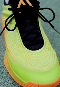 Limegrøn sneaker med en tekstureret overflade, svunget design, sort mesh tungespids, sorte snørebånd og orange detaljer på sålen.