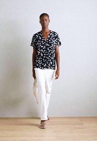 Top nero a maniche corte con motivo astratto bianco, design a V; abbinato a pantaloni bianchi e sandali chiari, con una borsa beige.
