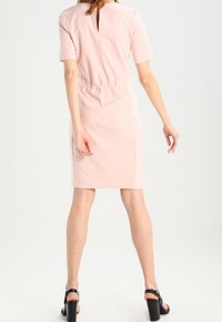 Vestido rosa ajustado con mangas cortas, detalle de ojal en la espalda y cintura elástica. Lleva sandalias negras de tacón, con textura suave.