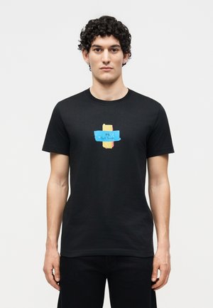 Bărbat cu păr creț purtând un tricou negru cu un grafic colorat în stil panglică, pe fundal simplu, cu inscripția „PS Paul Smith”.