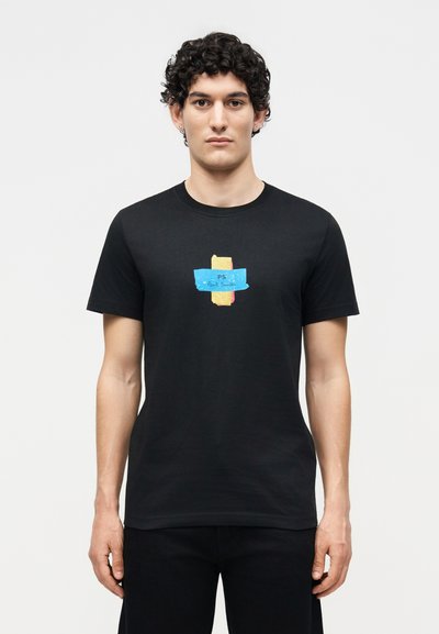 Άνδρας με σγουρά μαλλιά που φοράει μαύρο T-shirt με πολύχρωμο γραφικό σε στυλ ταινίας που γράφει "PS Paul Smith" σε μονόχρωμο φόντο.