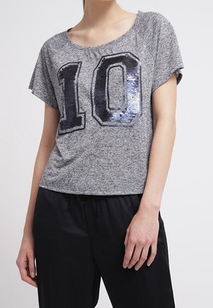 Kobieta ubrana w szarą koszulkę z krótkim rękawem z dużą cekinową cyfrą "10" i czarne spodnie, stoi z jedną ręką rozluźnioną.