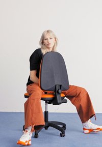 Black office chair with an orange seat and accents; features a round backrest and adjustable mechanism. Person seated, wearing casual clothes.