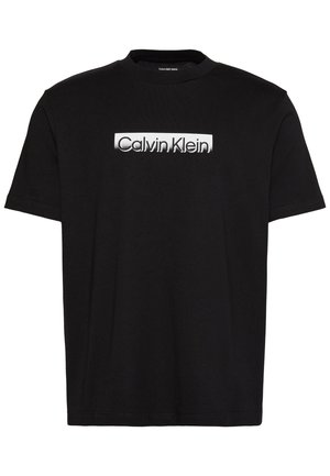 Μαύρο t-shirt με κοντά μανίκια και λευκή ορθογώνια ετικέτα στο στήθος που αναγράφει «Calvin Klein» με μαύρα γράμματα.