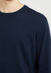 Granatowy sweter z okrągłym dekoltem wykonany z miękkiego materiału, z subtelnym logo na piersi oraz prążkowanymi detalami na dekolcie i mankietach.