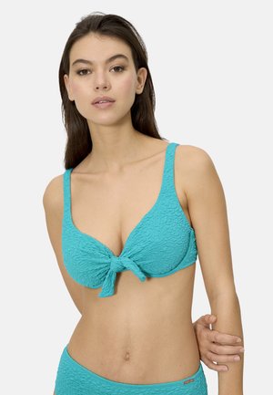 Joven mujer con top de bikini turquesa texturizado con nudo delantero, de pie frente a un fondo liso y claro, mirando hacia adelante.