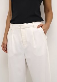 Pantalons blancs ajustés, avec des plis, un devant plat et des passants de ceinture. Associés à un haut noir sans manches. Texture lisse. Main reposant dans la poche.