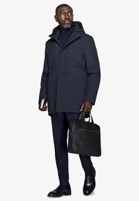 Marine vandtæt frakke med hætte, iført over en matchende turtleneck og ternede dressbukser. Sort læder buddeltaske og sko.
