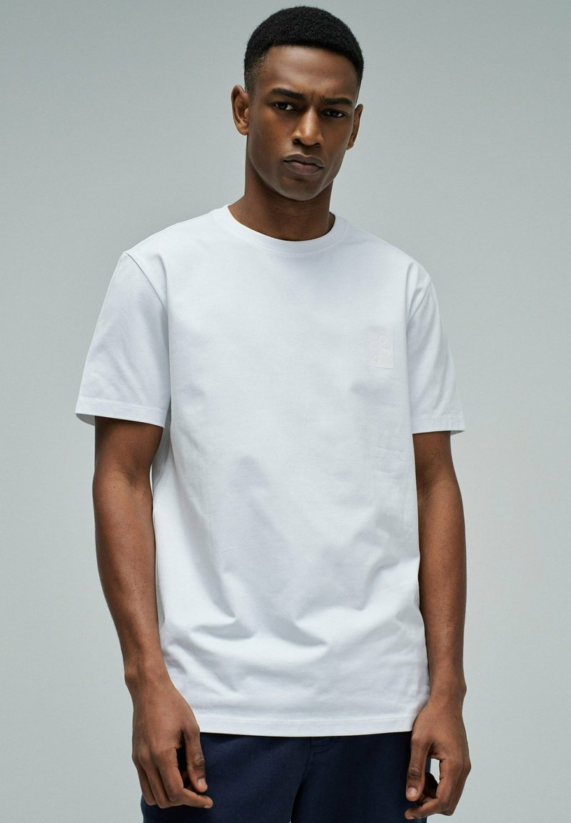 Joven con cabello corto que lleva una camiseta blanca de manga corta lisa y pantalones oscuros, posando frente a un fondo gris claro.