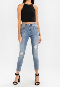 Jeans de mezclilla azules desgastados con acentos de piedras preciosas, ajuste slim y cintura alta. Combinados con un top corto negro de tirantes y tacones negros.