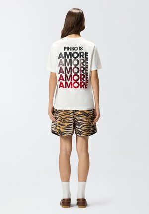 Donna con maglietta bianca con grande scritta multicolore "AMORE" e pantaloncini con stampa di tigre, in piedi di spalle su sfondo bianco uniforme.