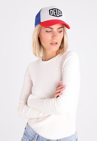 Gorra trucker roja y azul con un panel frontal blanco que presenta un logotipo negro de "DEUS", llevada con una camiseta de manga larga blanca con costillas.