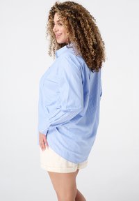 Chemise rayée bleu clair à manches longues et à coupe décontractée, avec une poche poitrine et des détails de poignet boutonné. Associée à un short crème.