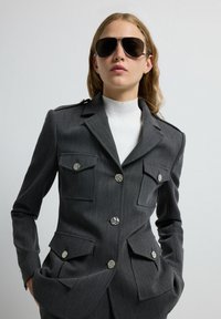 Blazer gris ajustado con dos bolsillos en el pecho, botones plateados y epaulettes en los hombros. Se lleva sobre un cuello de tortuga blanco con relieve. Modelo con gafas de sol oscuras.