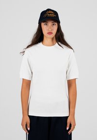 Biała, prążkowana koszulka z krótkim rękawem i okrągłym dekoltem, w połączeniu z ciemnymi spodniami. Granatowa czapka z pomarańczowymi wyszywanymi tekstami i detalami.