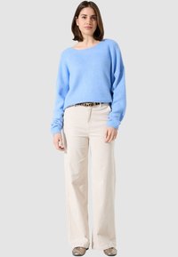 Un pull bleu clair à la coupe oversize avec des poignets côtelés, associé à un pantalon beige à jambes larges et une ceinture à motifs. Texture lisse et style décontracté.