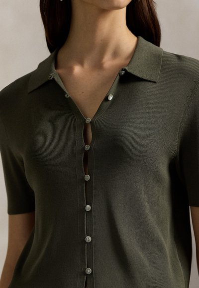Πράσινο λαδί κοντομάνικο πλεκτό τοπ με V-λαιμόκοψη, ριμπ ύφανση και μικρά κουμπιά μπροστά, που αναδεικνύει μια εφαρμοστή σιλουέτα.